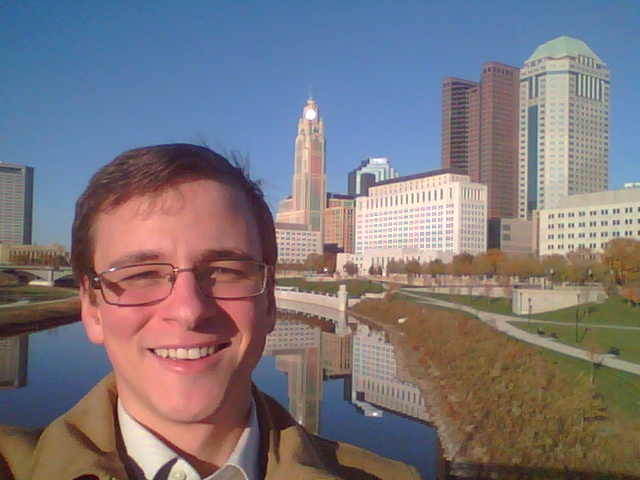 A photo of Grayson Hart standing in front of a city skyline with a lake seen in the distance. 