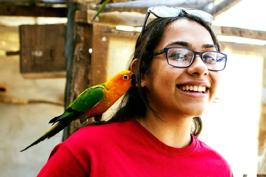 A photo of a lady smiling while the bird on her shoulder tries to nibble at her ear. 
